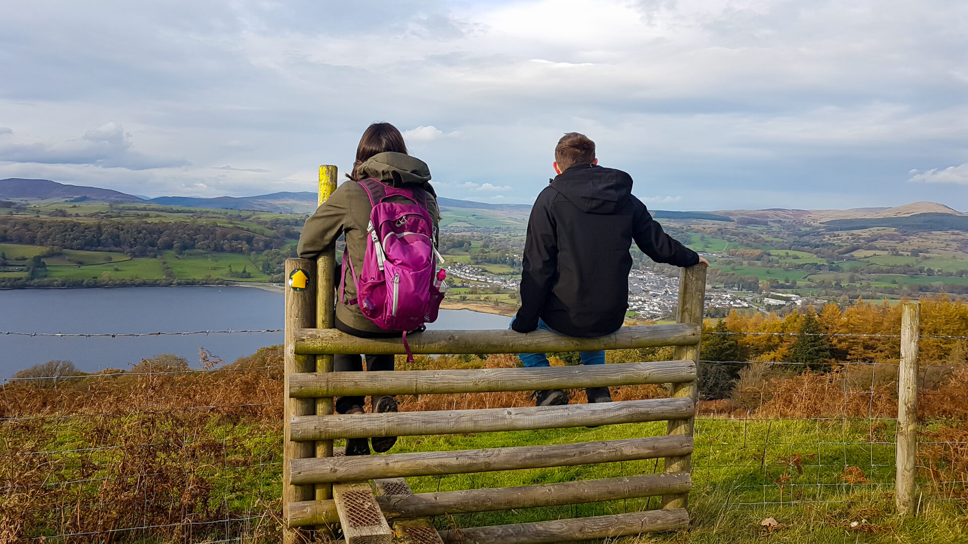 Two walkers sit on a wooden fence looking out at the scenery around them