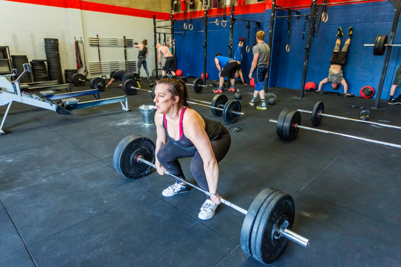 Woman lifting barbell in a deadlift at the gym, with other gym goers working our behind her.