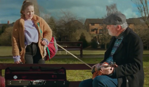 A woman approaching a park bench where a man is sitting holding a violin.