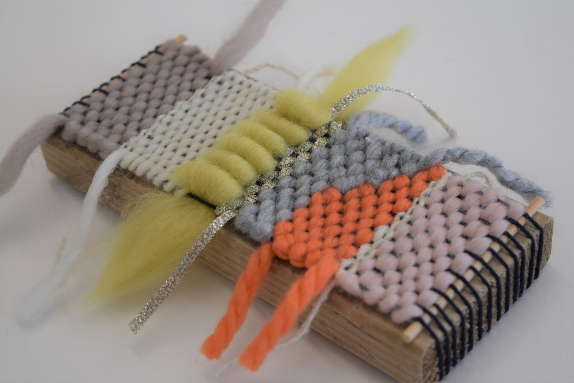 A small brick of wood with different coloured strands of yarn woven in (grey, white, yellow, orange and orange).