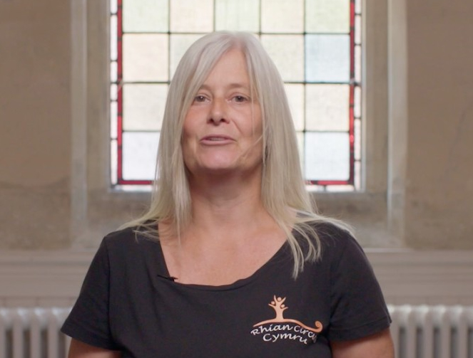 A head and shoulders shot of a person with long blond hair. They're looking into the camera and wearing a black shirt that says 'Rhian Circus Cymru'.