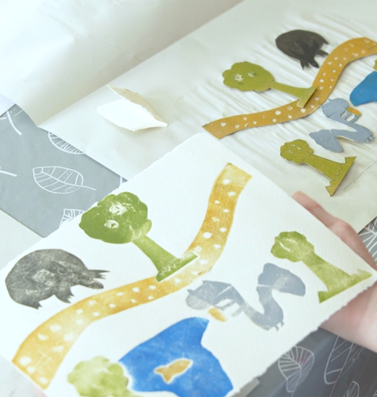 A piece of paper with different prints stamped onto it: tree, yellow path, lake, yellow fish and squirrel. Next to it, on another sheet of paper, are the cutouts that match the prints.