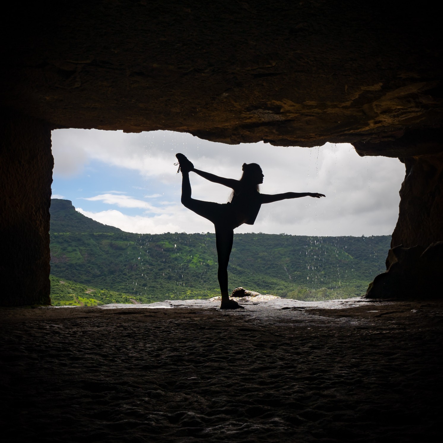 A silhouette of a person standing on one foot in front of a cave. One arm is outstretched, and the other is reaching back to grip their foot, which is held high in the air. In the background are green hills.