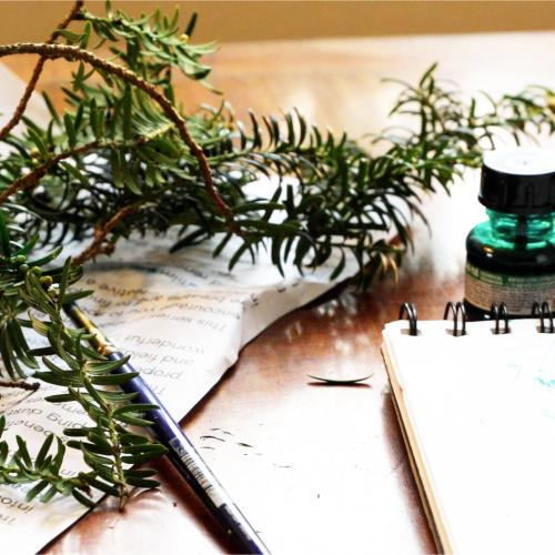 On a small table, several items are scattered: A small tree branch on a crumpled piece of paper; a small bottle of ink; a paintbrush; a sketch pad.