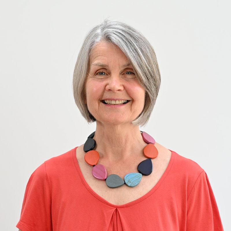 A head and shoulders shot of a smiling person with shoulder-length grey hair. They're wearing a colourful, large wooden necklace and a cotton coral top.