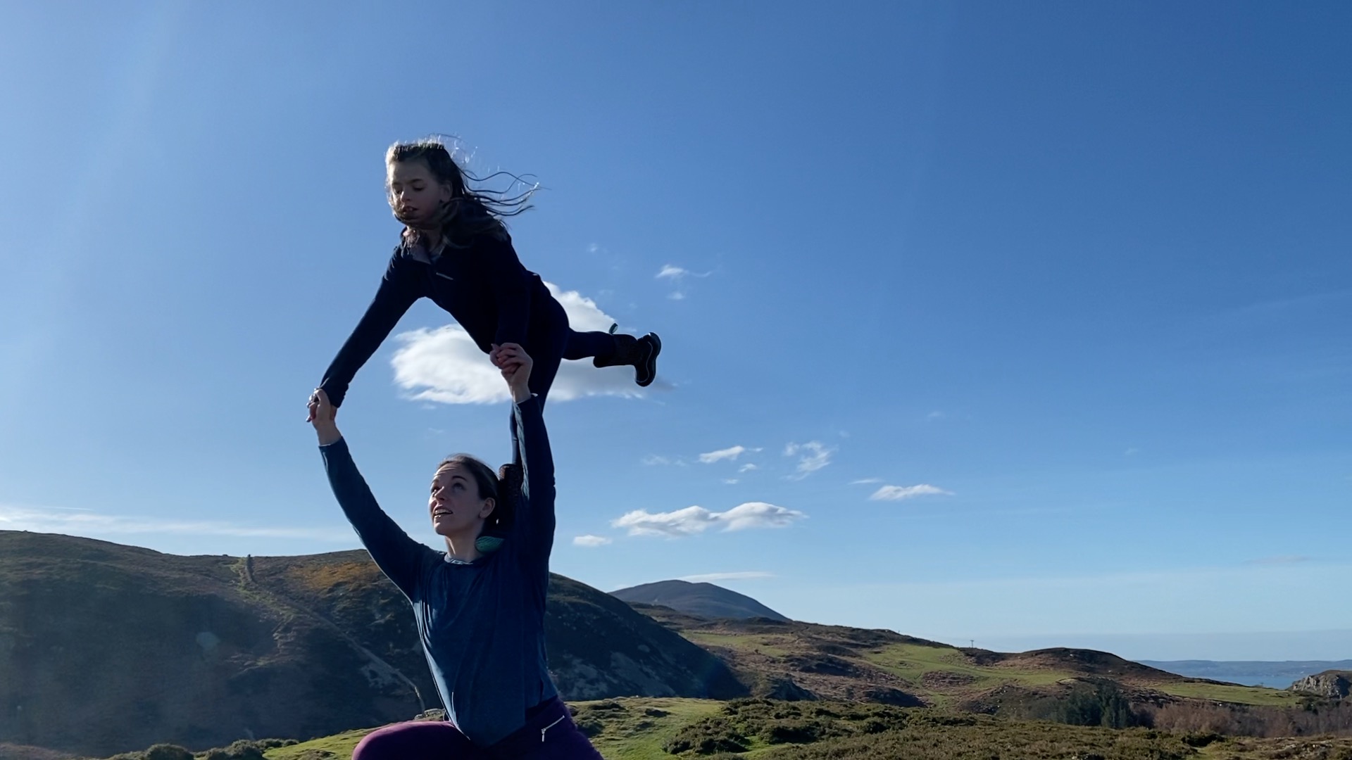 A person holds a child, who's standing on one leg on the person's left shoulder. The person is reaching up and holding the child's arms. In the background is a mountain range and bright blue sky.