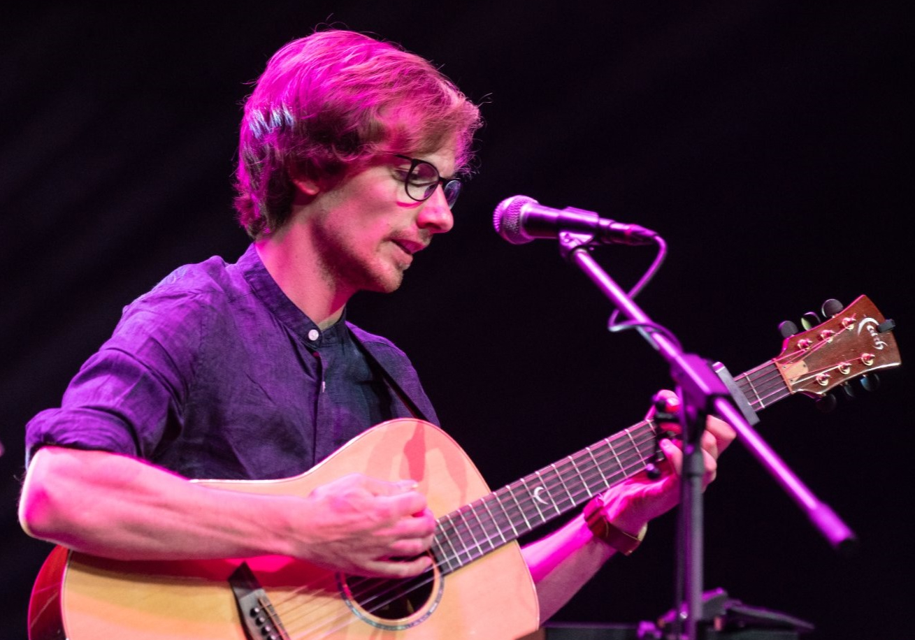 A person with short hair and glasses strums an acoustic guitar on stage. They're bathed in a bright magenta light and stand at a microphone.