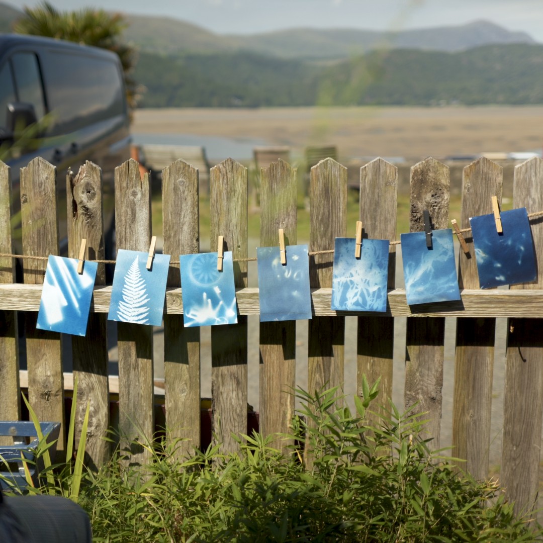 Seven postcards with blue and white patterns are pinned to a long string that's attached to a wooden fence. Mountains are in the background.