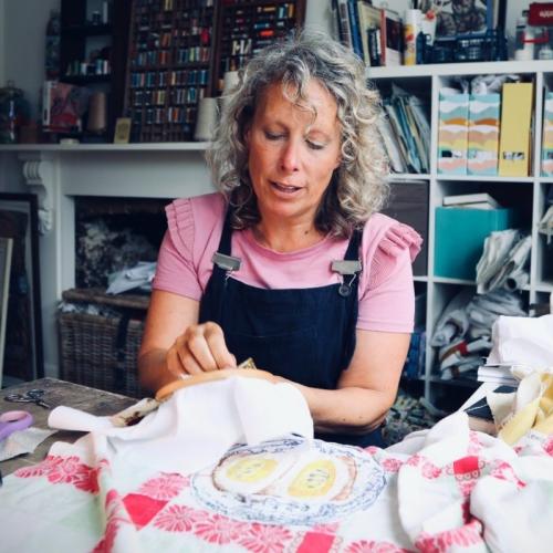 Person with shoulder-length curly blond hair sits at a table in a studio. They're holding an embroidery ring and adding a stitch to it. Different types of textiles are scattered around the table.