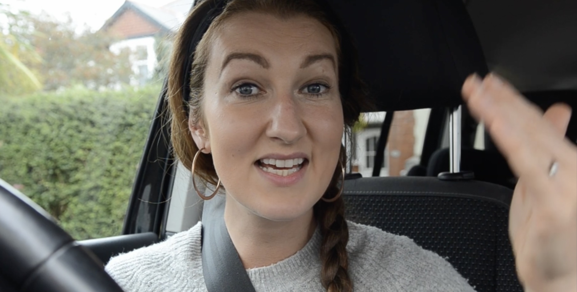 Head and shoulders shot of a person sitting in the driver's seat of a car. Their mouth is open (as if in mid-song) and their left hand is raised. They're wearing a grey jumper, hoop earrings and a black headband.