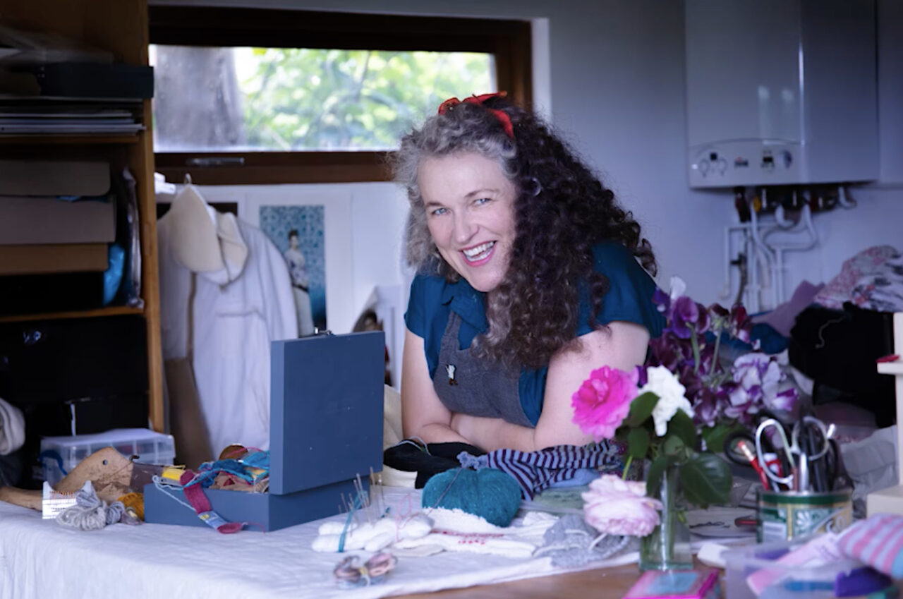 Smiling person with grey and brown curly hair crosses their arms and looks into the camera. They're sitting at a desk surrounded by sewing kits and materials.