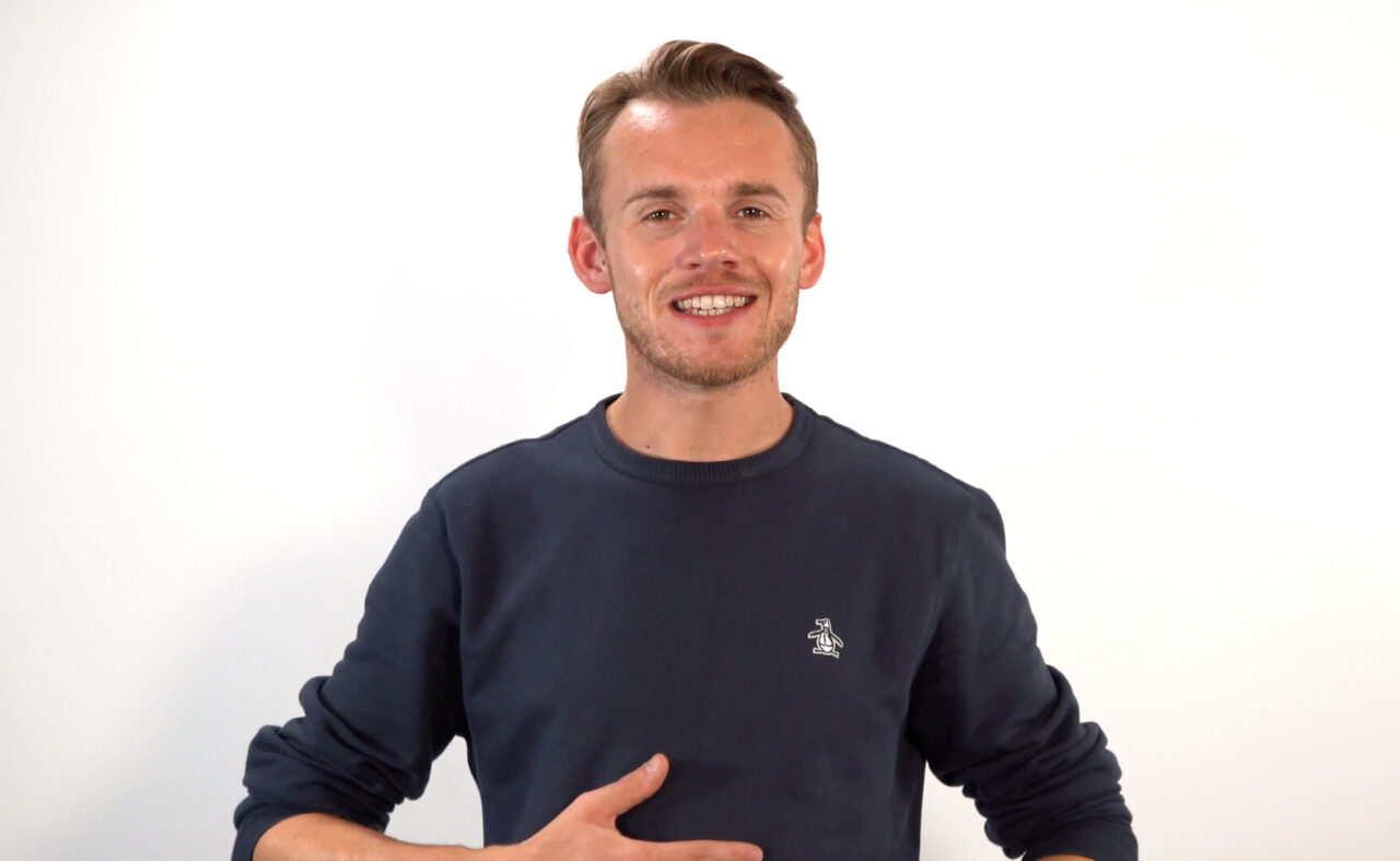 Person with short blond hair facing the camera with their hand resting on their torso. They're standing against a white background and wearing a long-sleeved navy top.