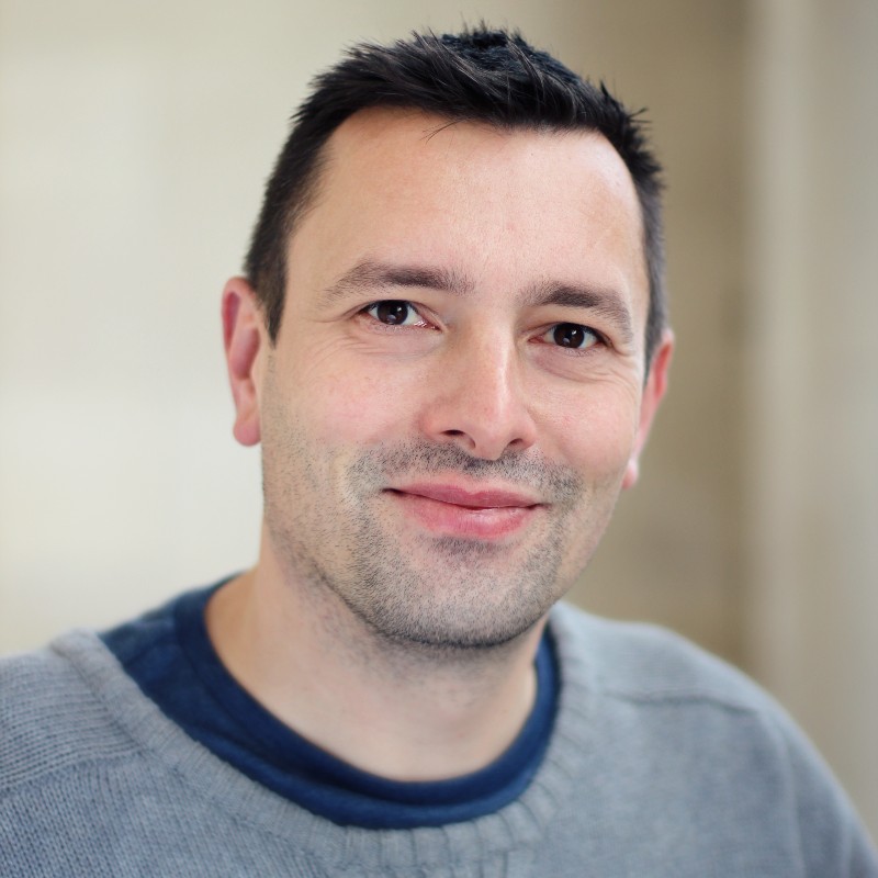 Portrait of a smiling person with short dark hair and dark eyes. They're standing against a beige background and wearing a grey jumper with a blue T-shirt underneath.