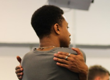 A head-and-shoulders shot of a person tightly hugging themselves. They're wearing a dark grey shirt and silver rings.