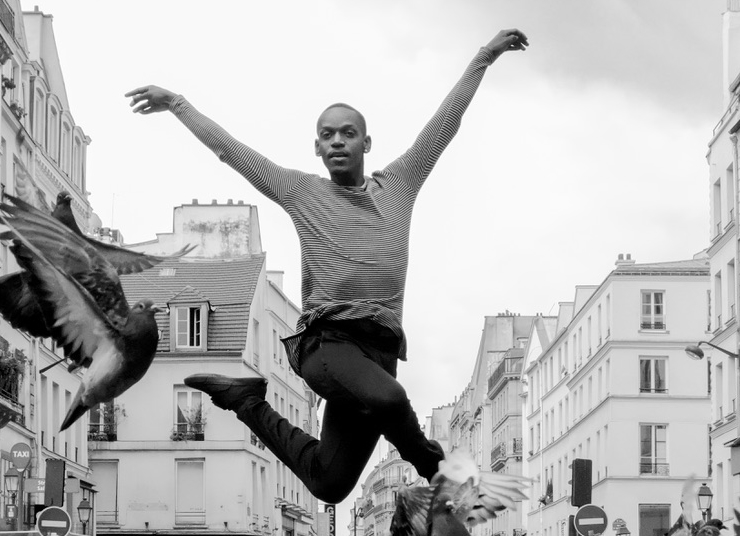 A black and white photo of a person jumping with their arms outstretched, mid-air, as pigeons flock around them. A city is in the background.