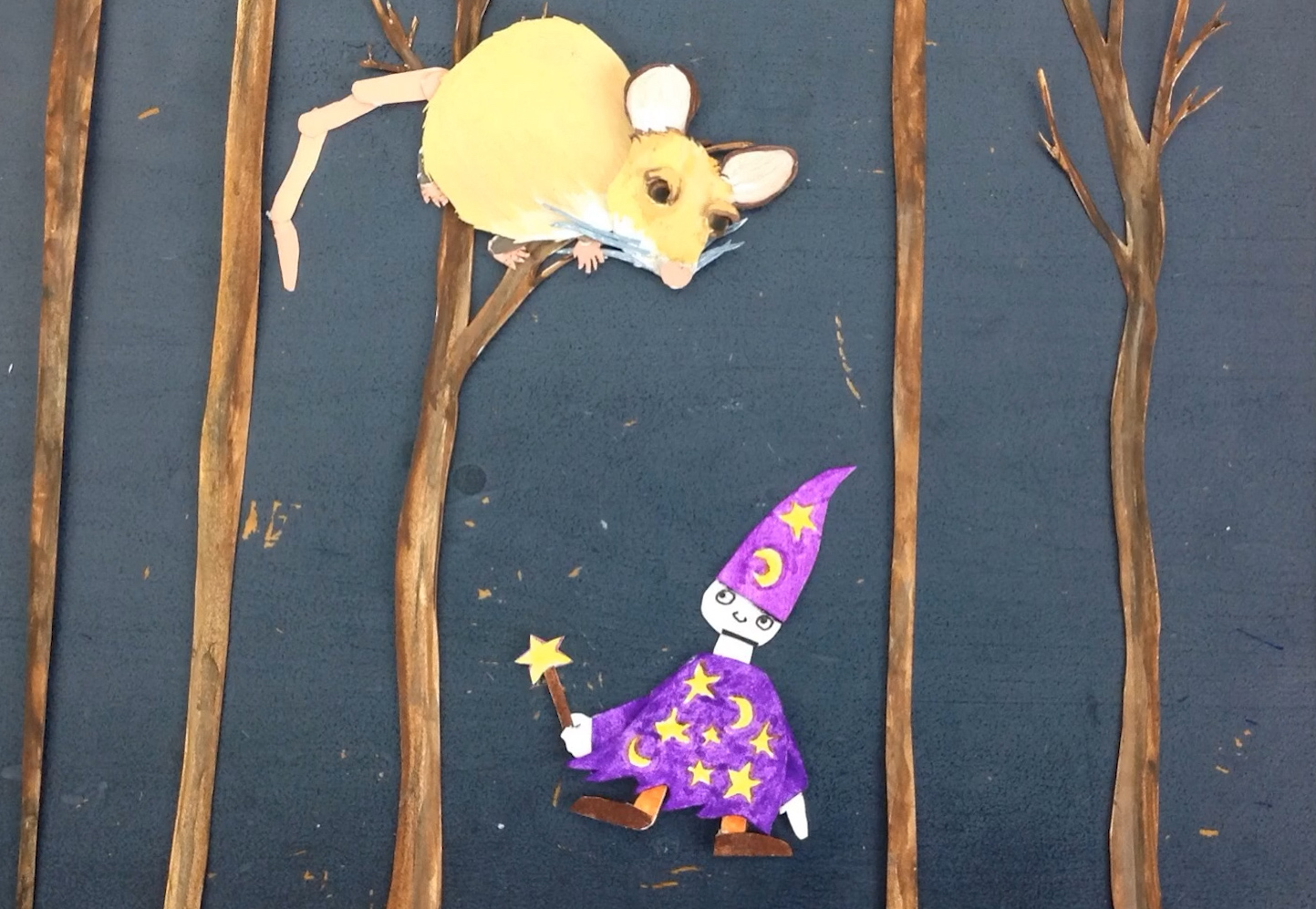 On a dark board, a cut-out drawing of a mouse sits atop a thin, painted tree. It's surrounded by four other thin trees. The mouse is looking down at a cut-out drawing of a smiling wizard in a purple hat and cloak with yellow stars and moons.
