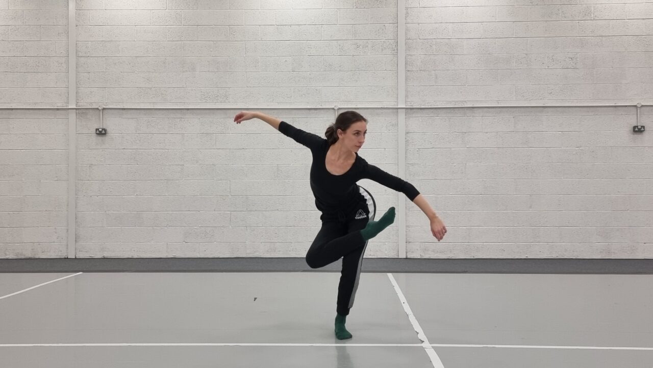 In a brightly lit dance studio, a person stands in a black top and sweatpants with their arms outstretched, bending their body and looking off camera. Their right leg is crossed over their left knee.