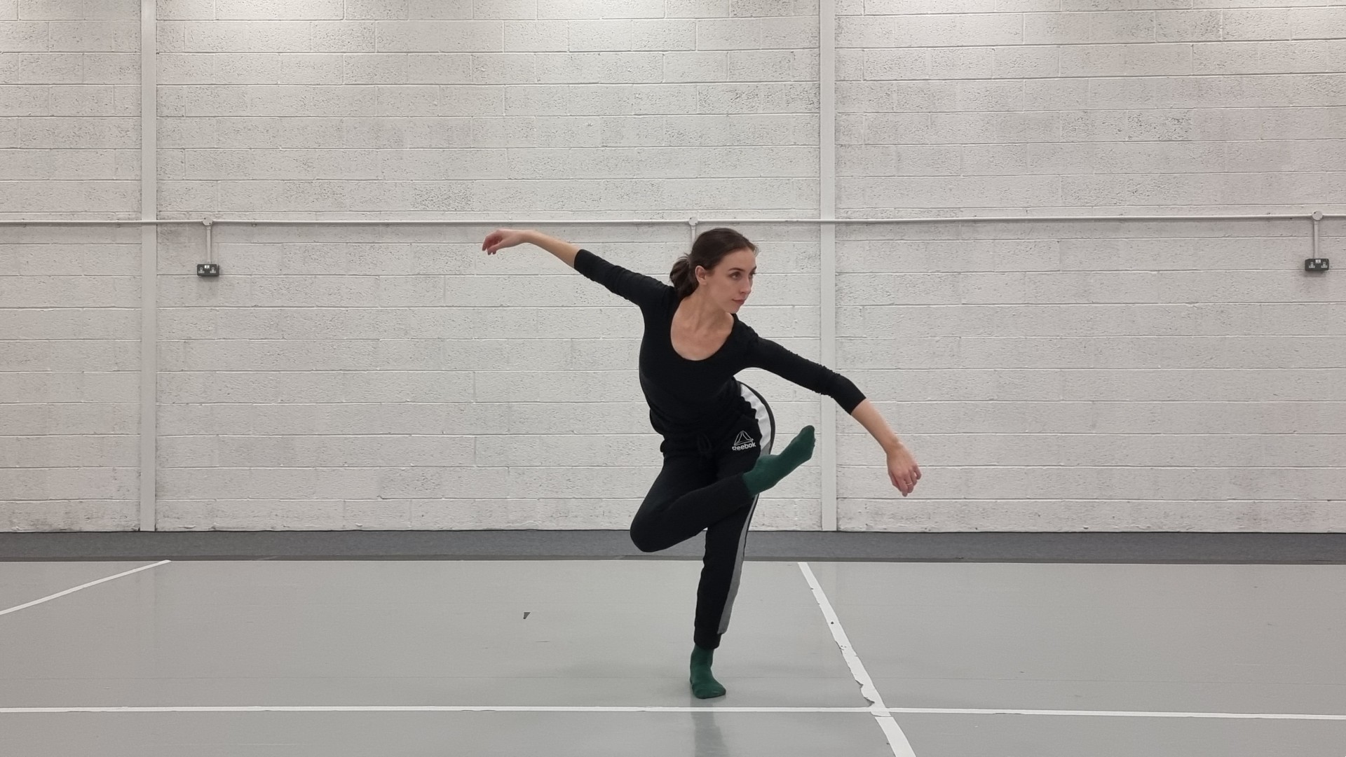 In a brightly lit dance studio, a person stands in a black top and sweatpants with their arms outstretched, bending their body and looking off camera. Their right leg is crossed over their left knee.