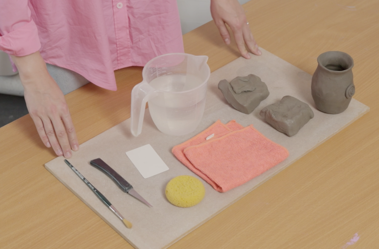 Fingertips touch a board with a variety of materials on it: plastic water jug, half-full; a folded pink cloth; a small, round yellow sponge; two squares of grey clay; a tiny vase fashioned out of grey clay; a paint brush; small square sheet of paper; potter's knife. The board is sitting on a light wooden table. The person is wearing a pink cotton shirt.