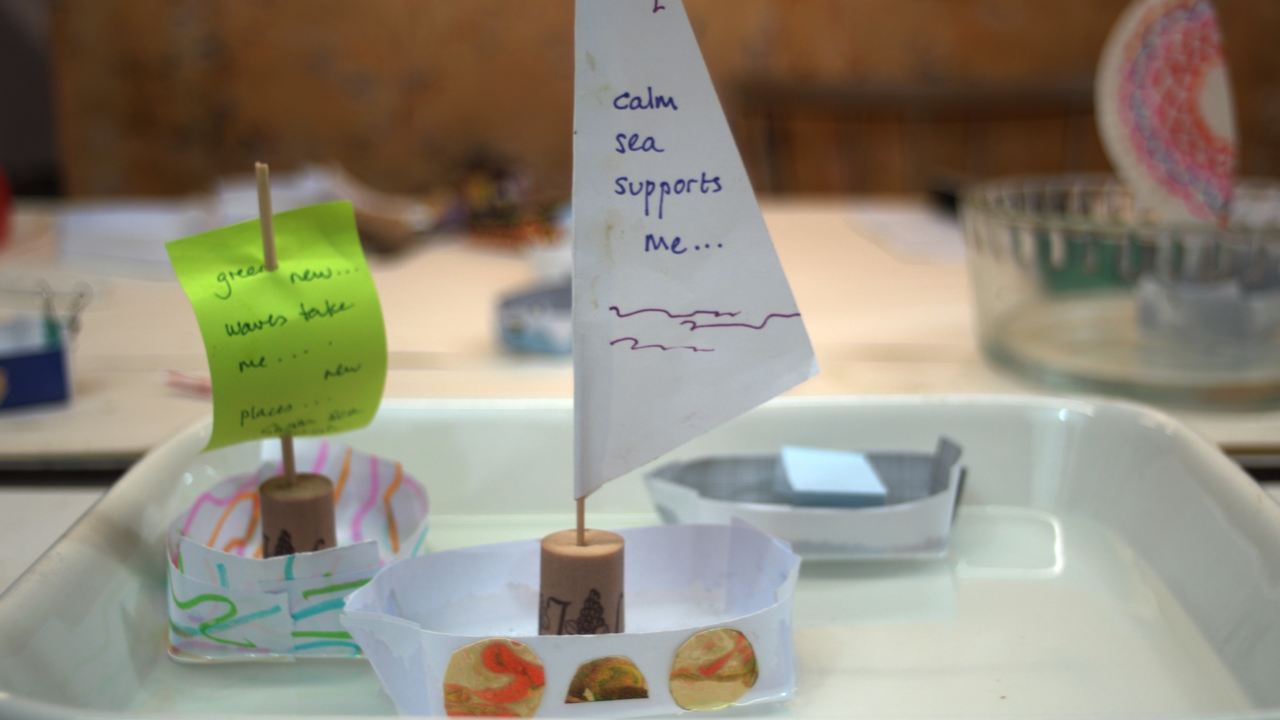Three paper boats floating in a ceramic roasting dish filled with water. One boat has a green Post-It as its sail, with the words 'green new... waves... take me... new places...' written in marker. The other boat has a white paper mast with the words 'calm sea supports me...' written in on it in blue marker.