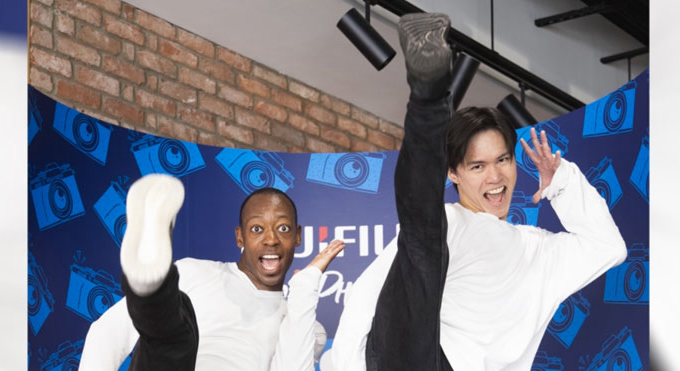 Two people standing in front of a blue and navy wall. They're wearing long-sleeve white tops, black trousers and trainers. They're kicking their right legs high into the air and looking joyfully into the camera.