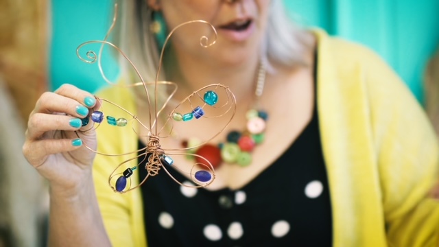 A person with blond hair holds up a butterfly fashioned out of copper wire. It's decorated with blue, navy and green beads.