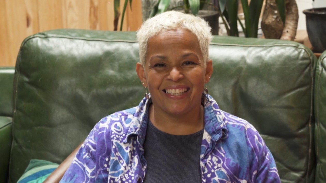 Photo of a smiling person with short blond hair sitting in a green arm chair. They're wearing a black T-shirt with a purple, blue and white shirt over it.