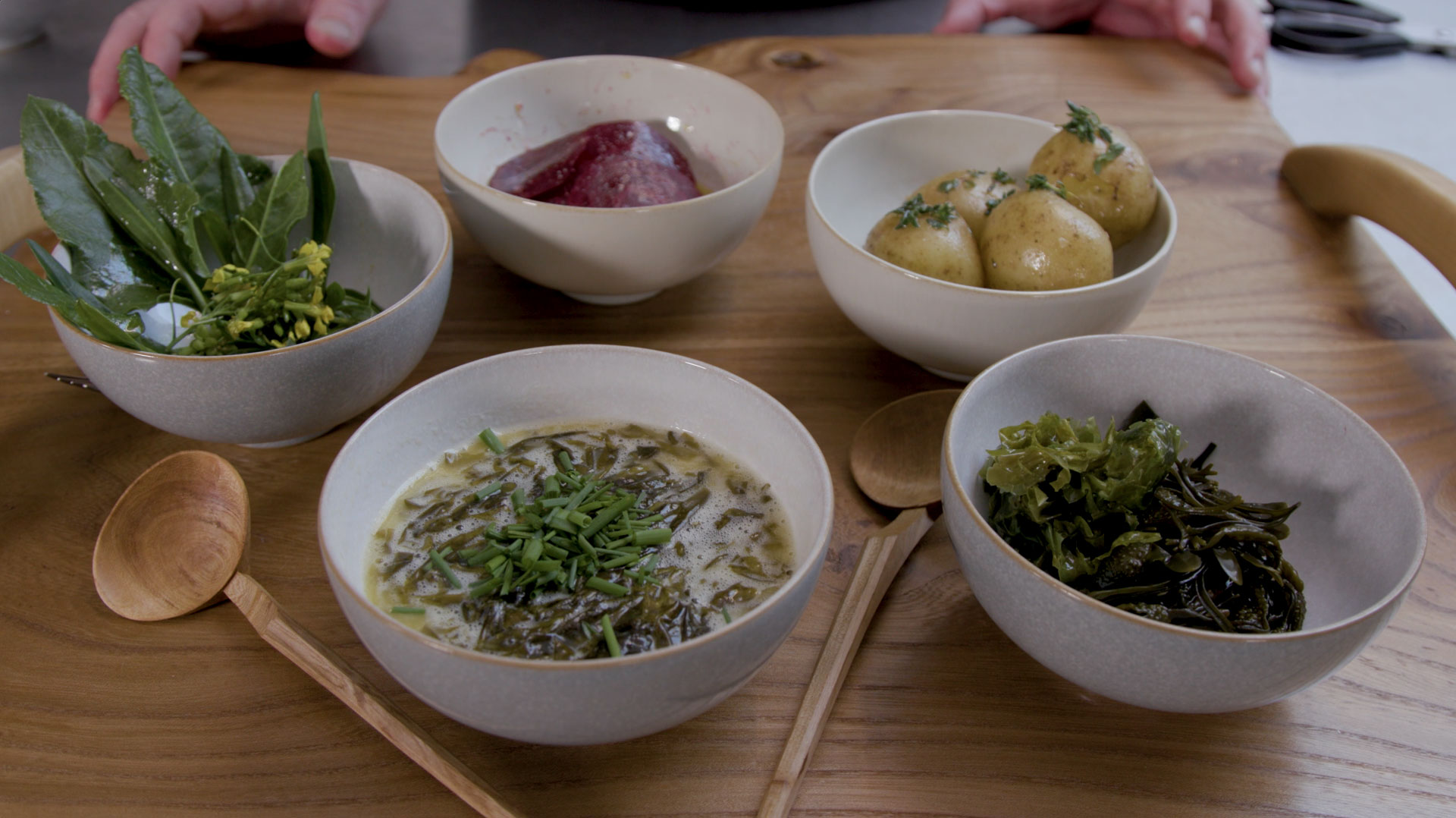 Five ceramic bowls sit on a light wooden tray. The bowls are filled with seaweed, soup, leaves, potatoes and a slices of beetroot. Two wooden spoons are lying next to the bowls.