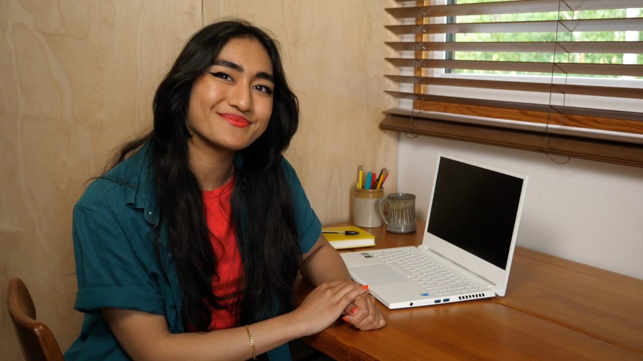 A person with long dark hair sits at a desk with a white laptop open. They're wearing a teal green shirt with a red shirt underneath. They're looking into the camera and smiling.