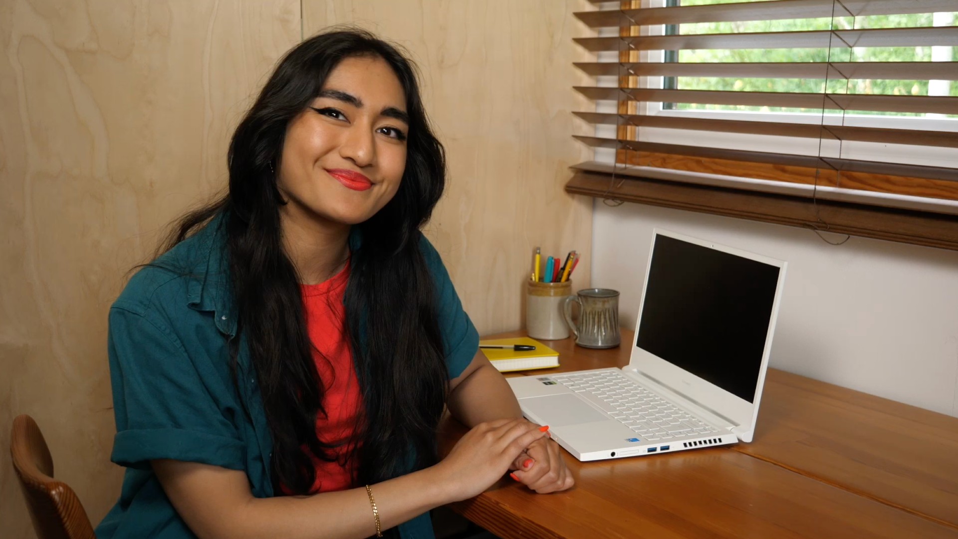 A person with long dark hair sits at a desk with a white laptop open. They're wearing a teal green shirt with a red shirt underneath. They're looking into the camera and smiling.