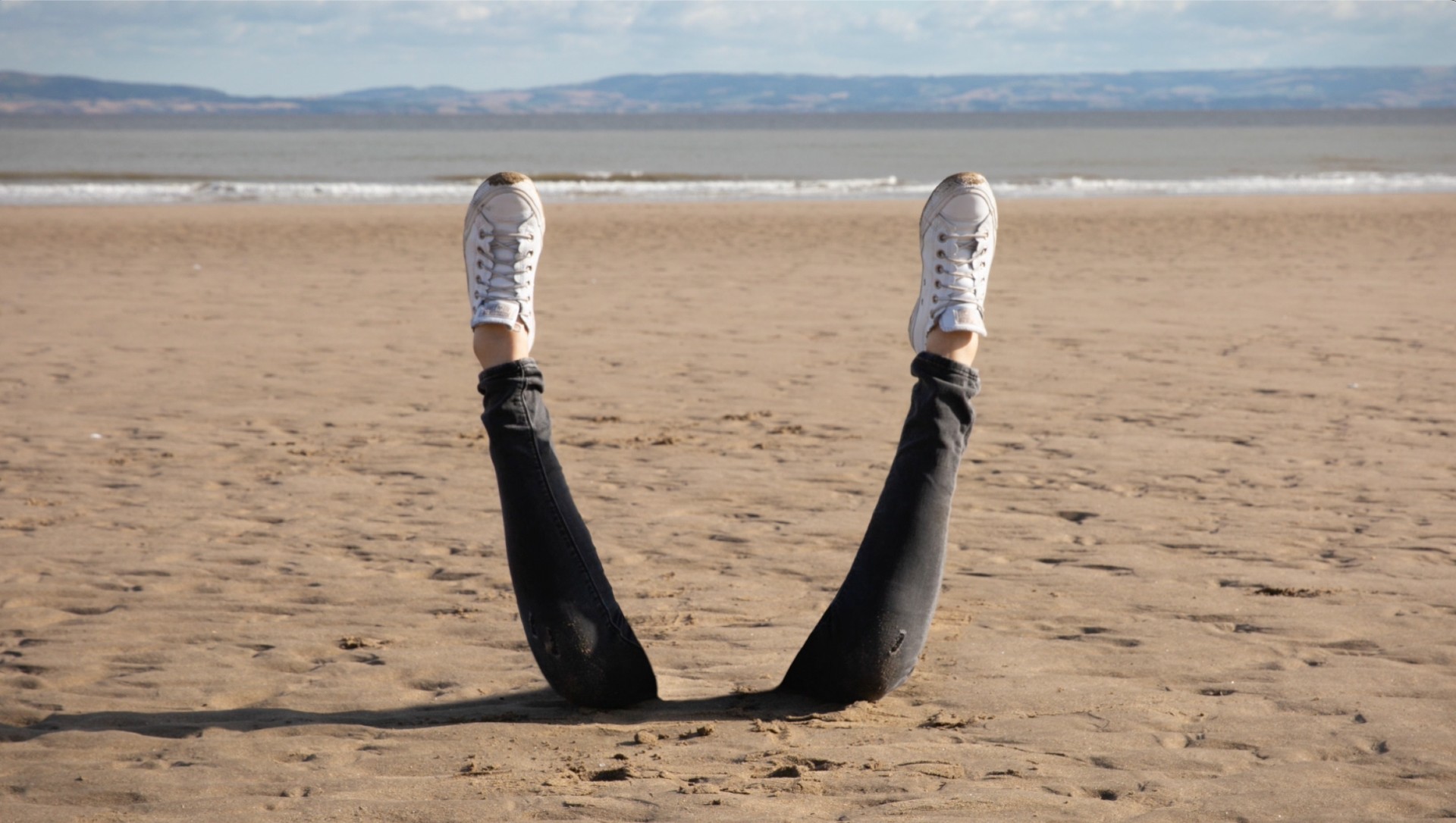 Two legs sticking out of the sand on a beach. The legs are clad in black jeans and white trainers. The ocean and a mountain range are in the background.