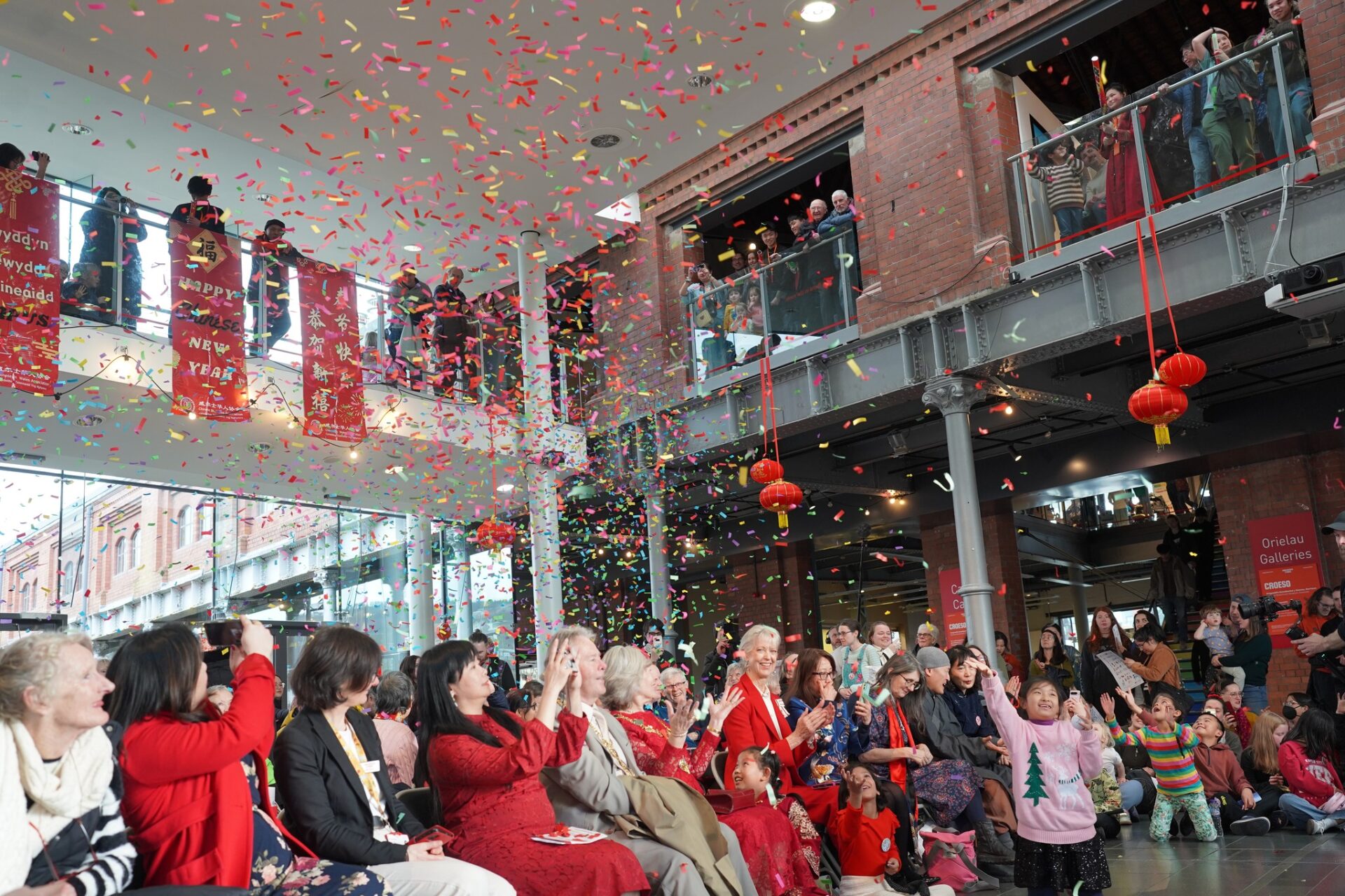 Crowd at an indoor celebration throws colorful confetti as banners and lanterns hang above.
