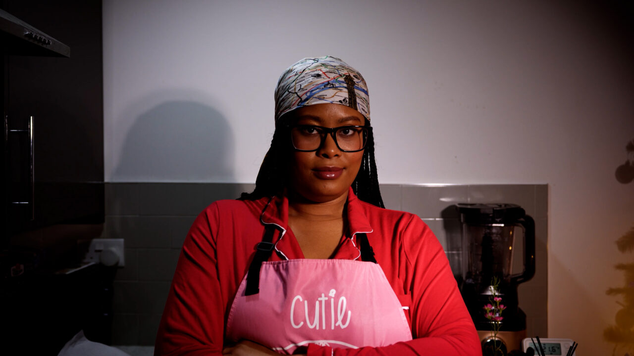 Woman wearing glasses and a patterned headscarf in a pink apron labeled 'Cutie' stands in a kitchen with arms crossed.