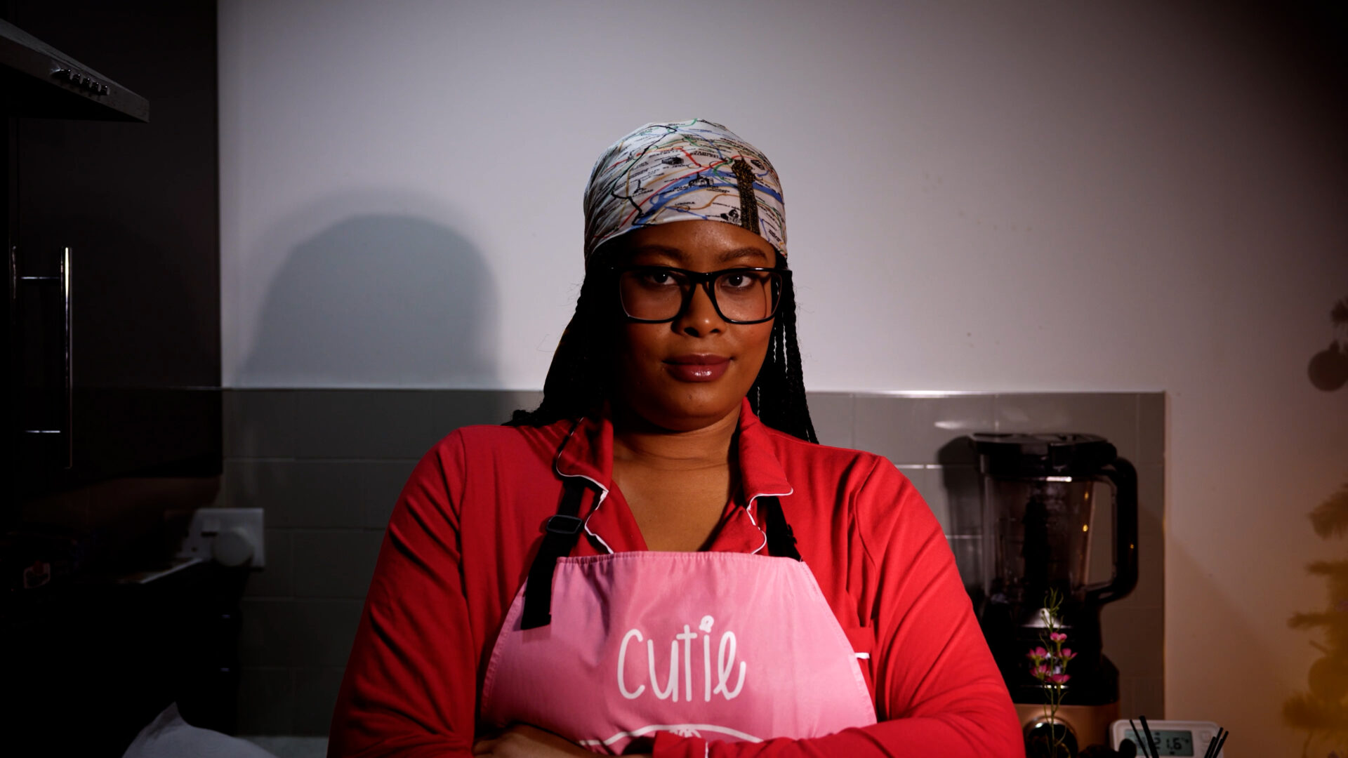 Woman wearing glasses and a patterned headscarf in a pink apron labeled 'Cutie' stands in a kitchen with arms crossed.
