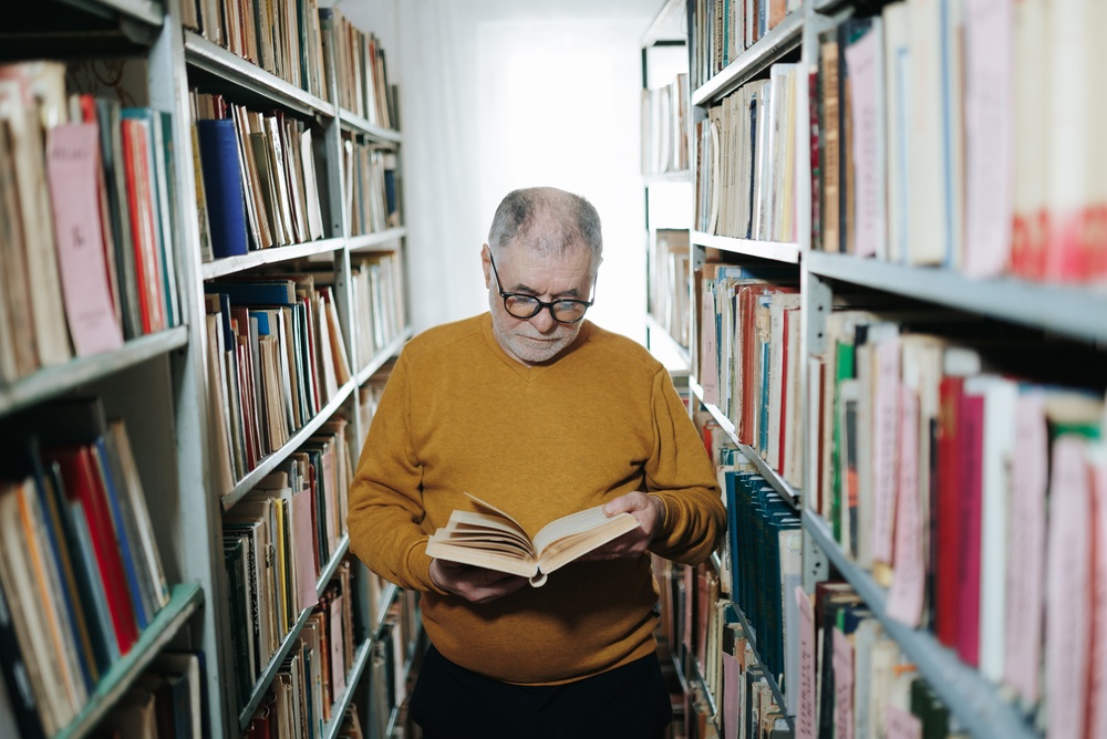 Older man with glasses reading a book in a narrow library aisle.
